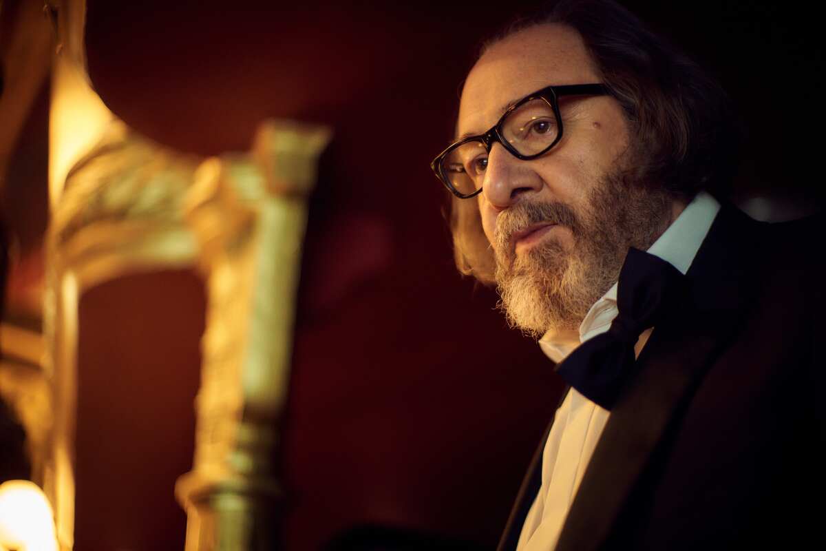 A man in a tuxedo, with bow tie open, and glasses sits in a gilded theater venue.