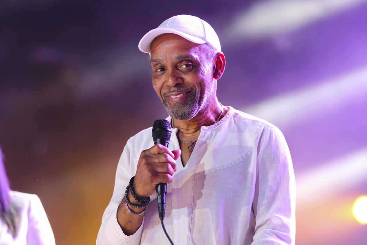 Frankie Beverly con sombrero blanco y camisa de lino sostiene un micrófono en el escenario