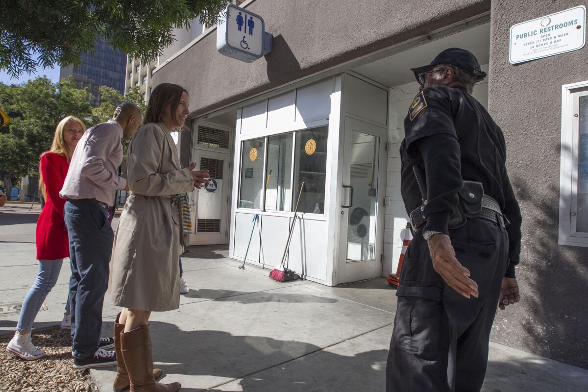Three people wait in a line on a sunny sidewalk outside a public restroom, as a woman in a security guard uniform looks on.