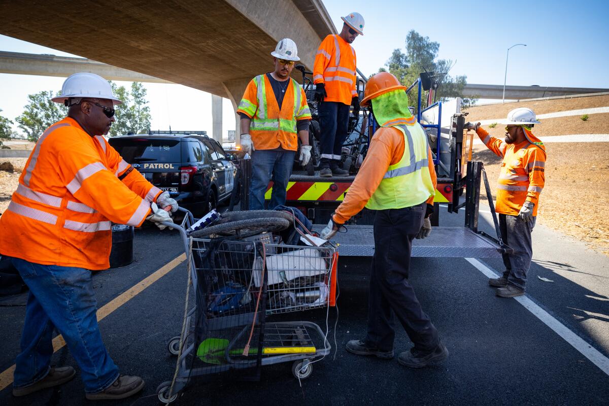 Workers in hard hats and high visibility clothing stand with a shopping cart on the shoulder of a road.