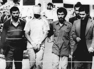 A blindfolded man, his hands bound, is flanked by other men in a black-and-white photo