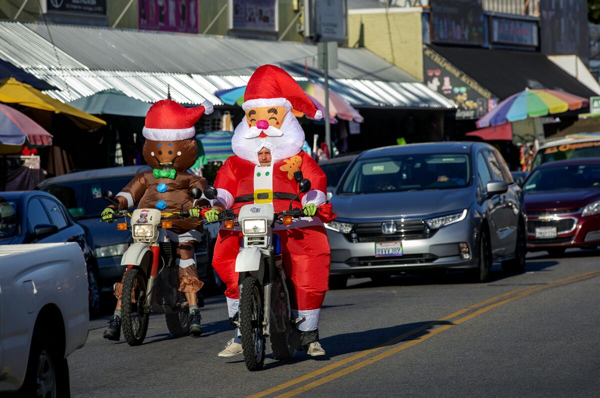 Two people ride motorcycles, one wearing a gingerbread man inflatable costume and the other wearing a Santa costume.