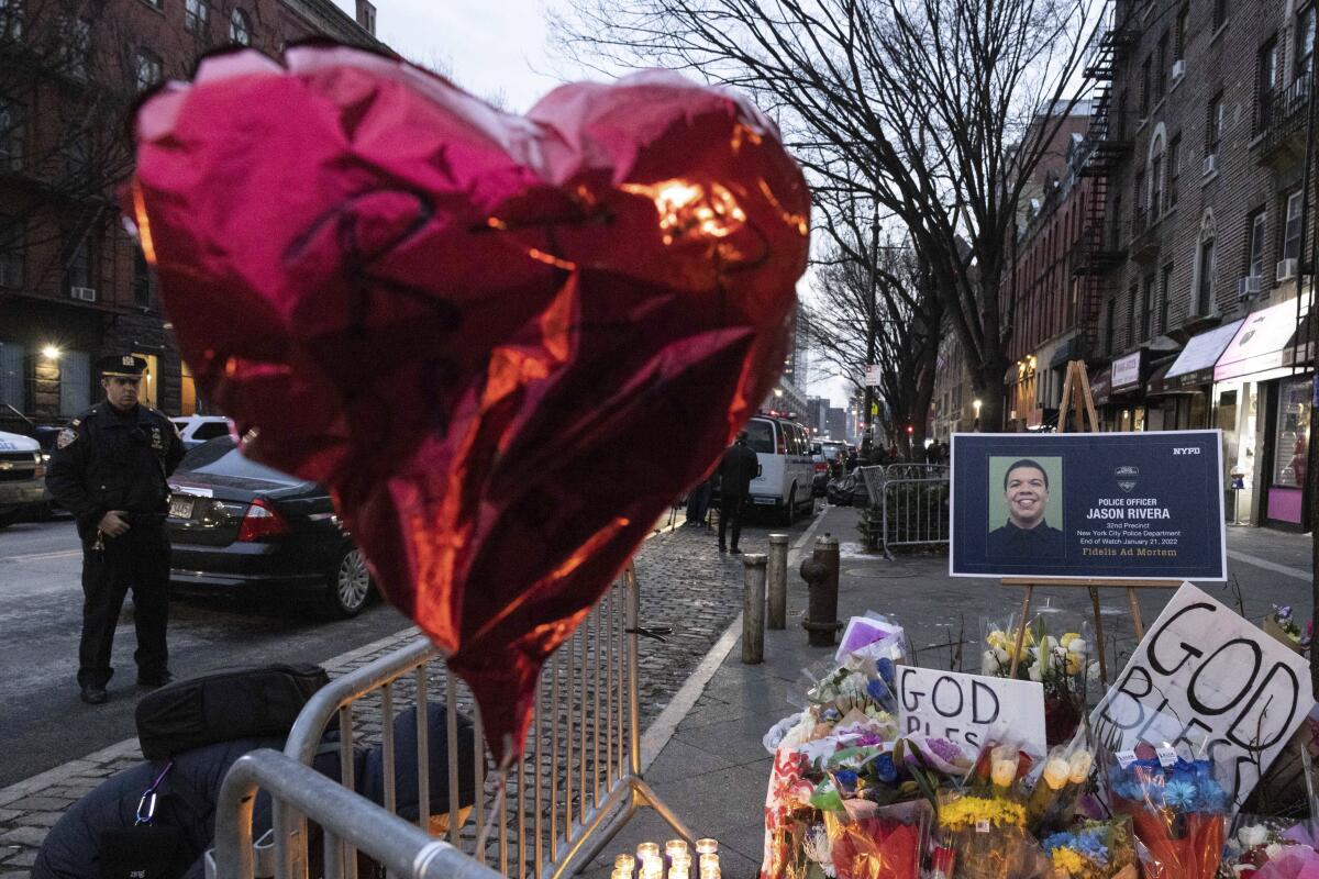 A sign with a photo of Officer Jason Rivera is surrounded by flowers and a red heart balloon on a sidewalk 