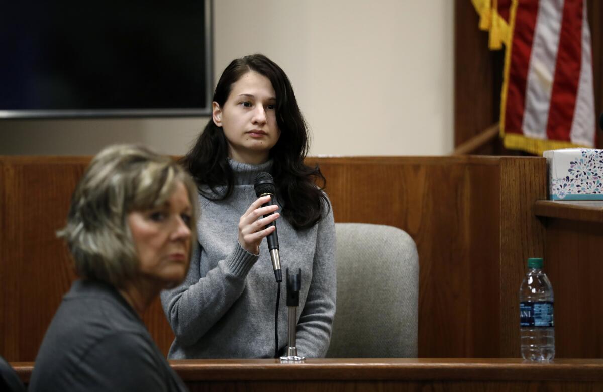 A woman with long brown hair in a gray sweater holding a microphone in a courtroom
