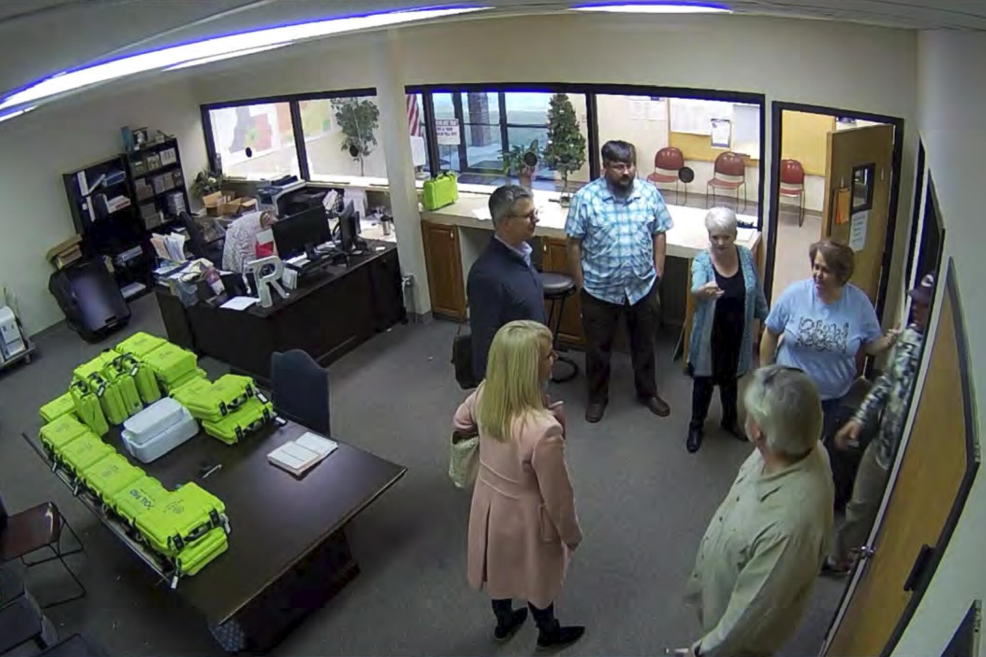 A view from a security camera of several people standing in an office near a table with voting equipment stacked on top.