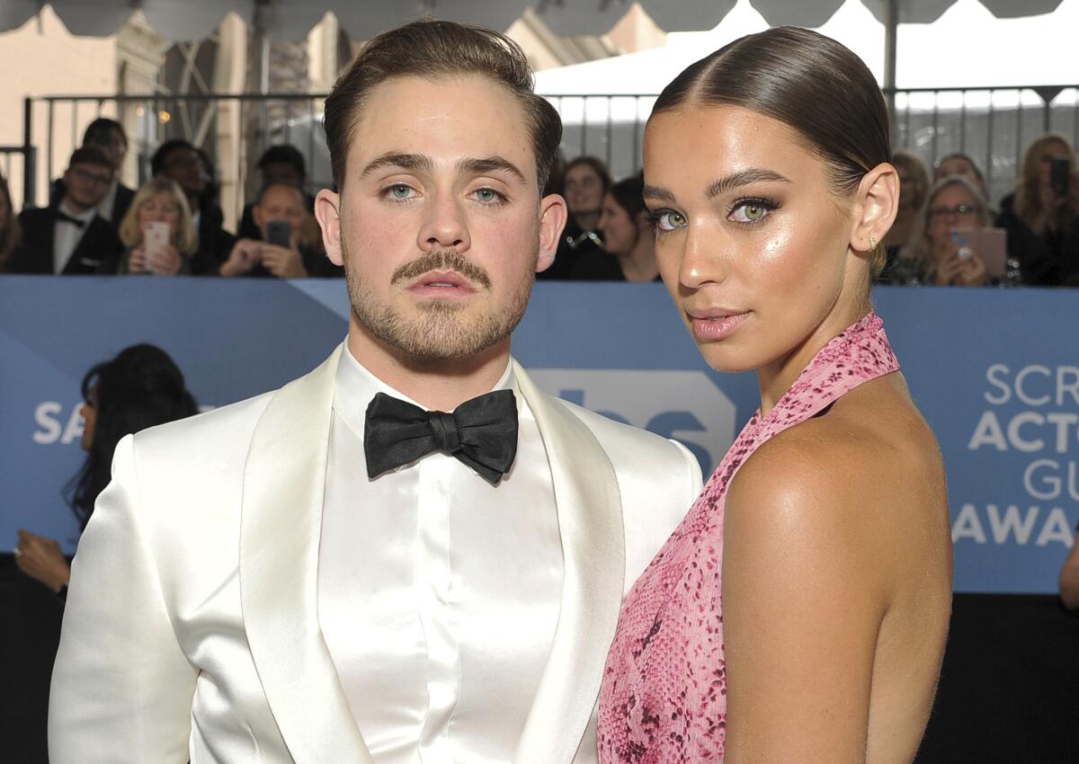 Dacre Montgomery in a white tuxedo standing next to girlfriend Liv Pollock in a pink, patterned halter dress