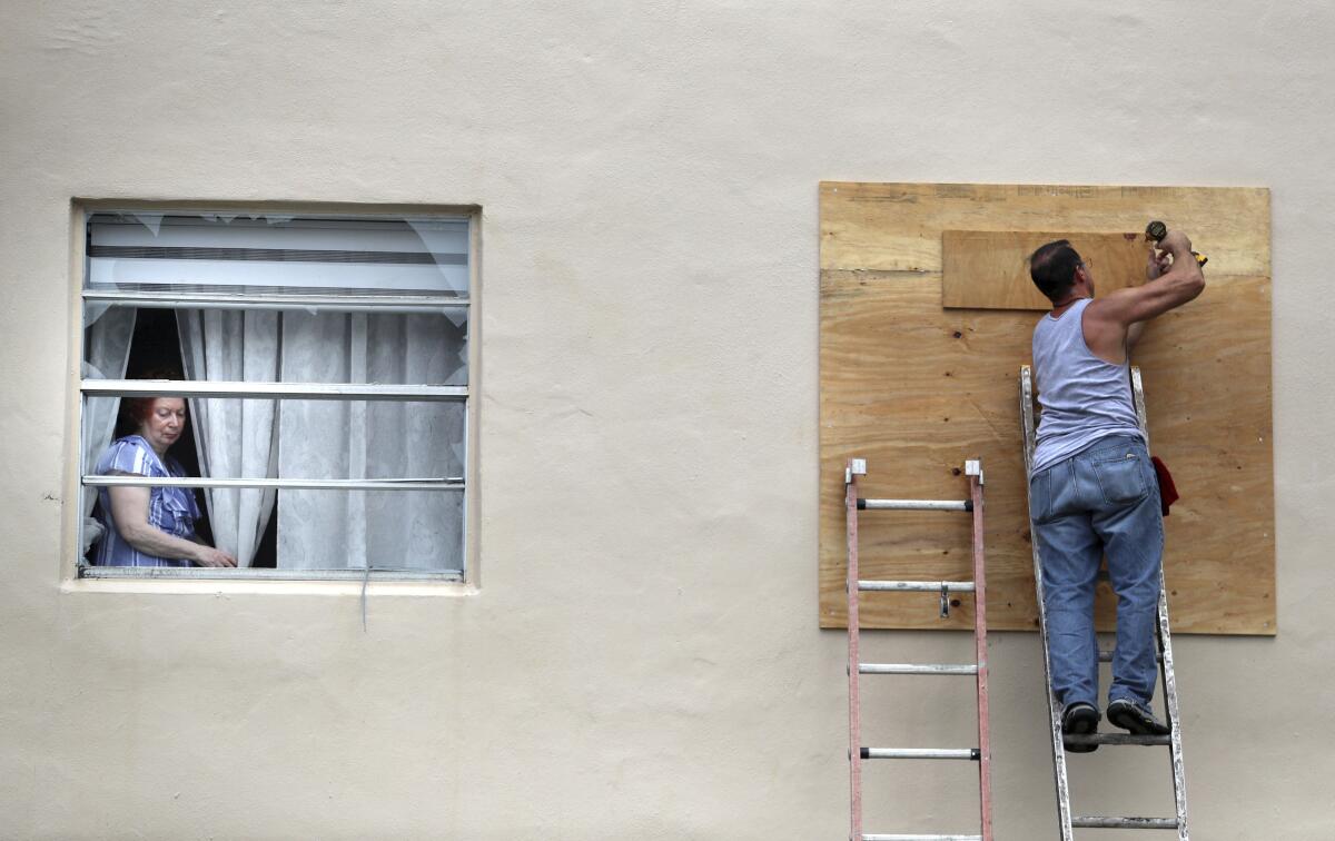 Una mujer se asoma por una ventana rota mientras un hombre tapia otra también dañada