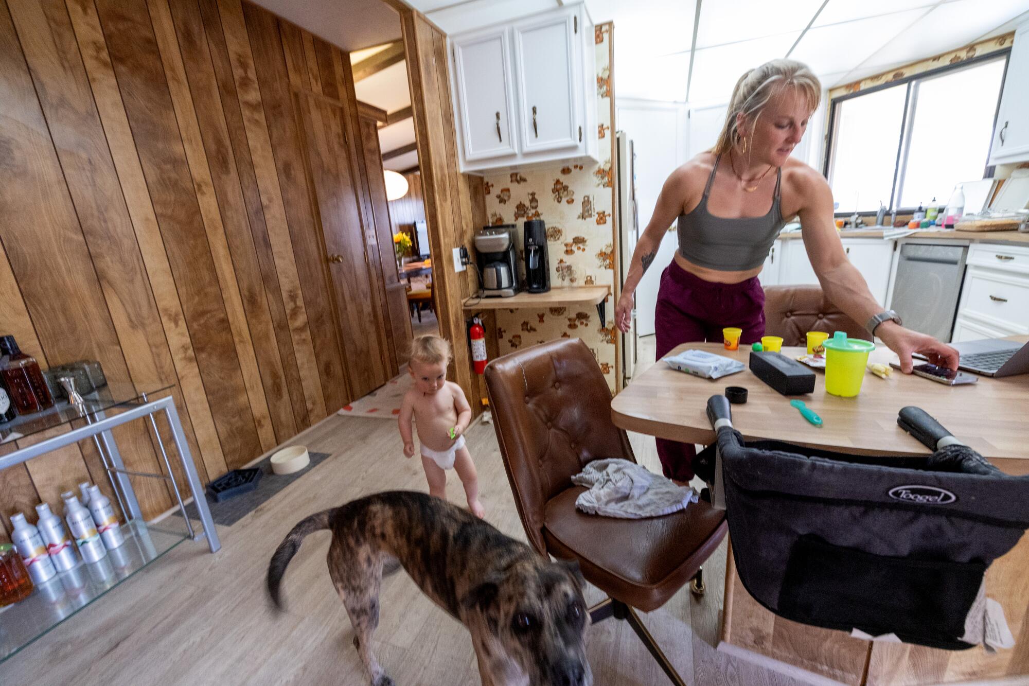 一位运动女士在厨房里和附近穿着尿布的幼儿一起查看手机。  
