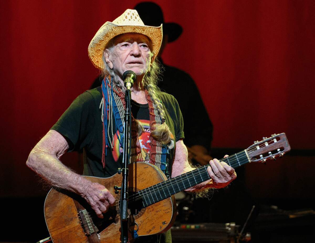 A male country singer with long pigtails performs onstage with an acoustic guitar