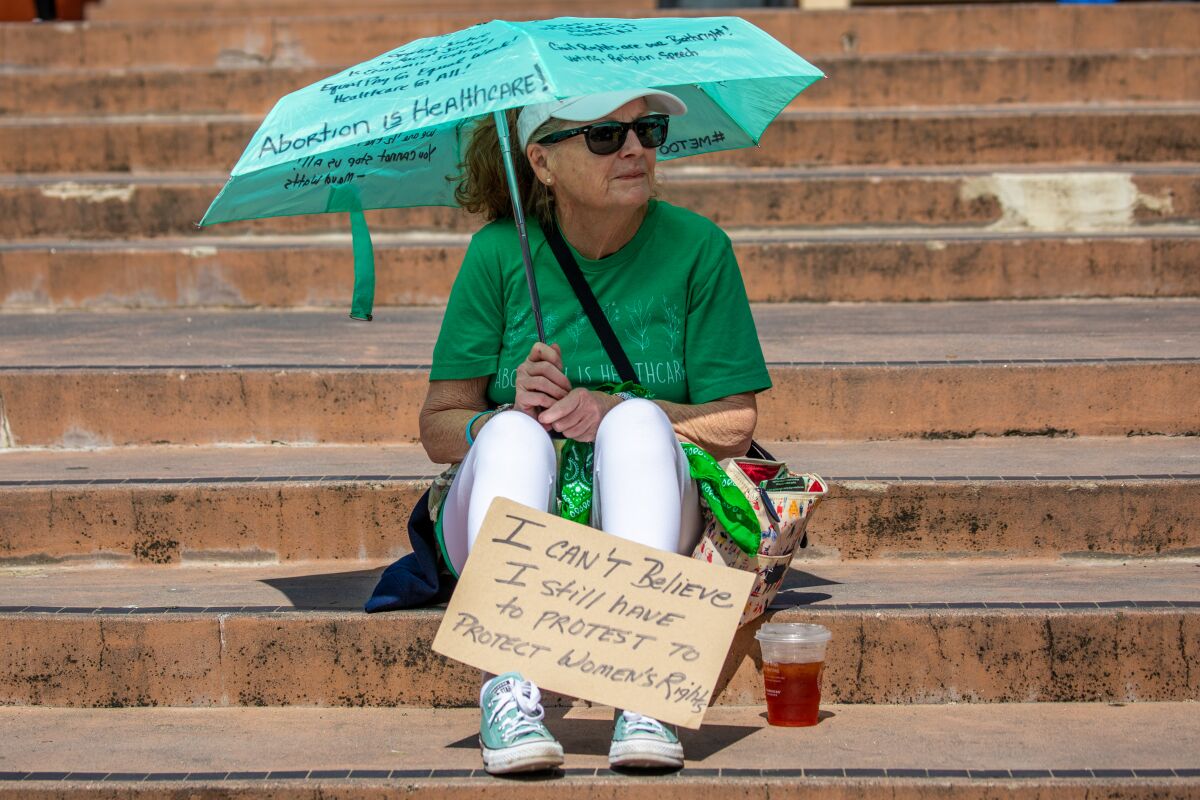 A woman sitting outside on steps with a handmade sign and an umbrella scrawled with reproductive rights messages in marker.