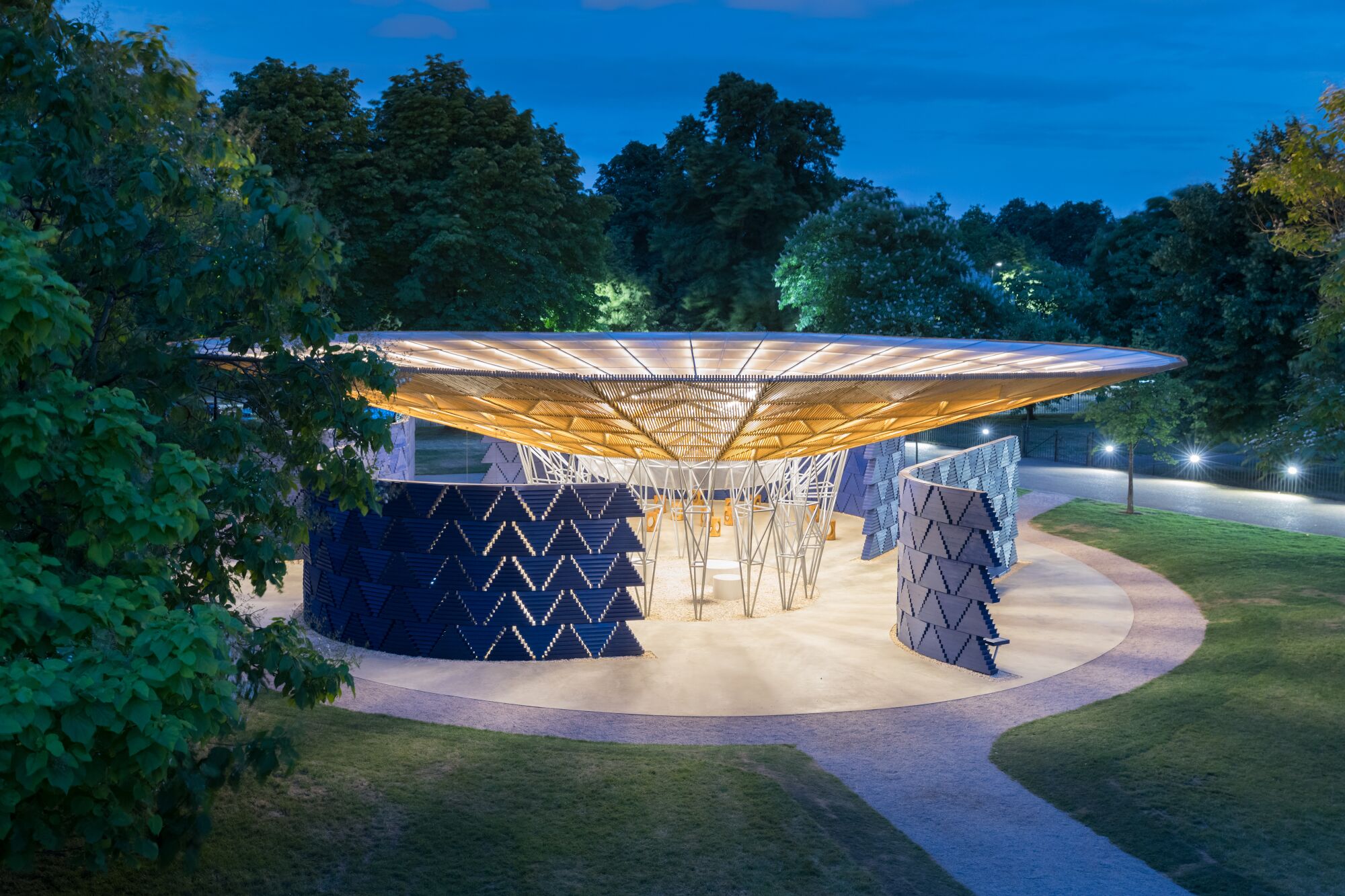 An image taken at dusk shows a pavilion with a circular roof over blue block walls punctured with a triangular pattern.