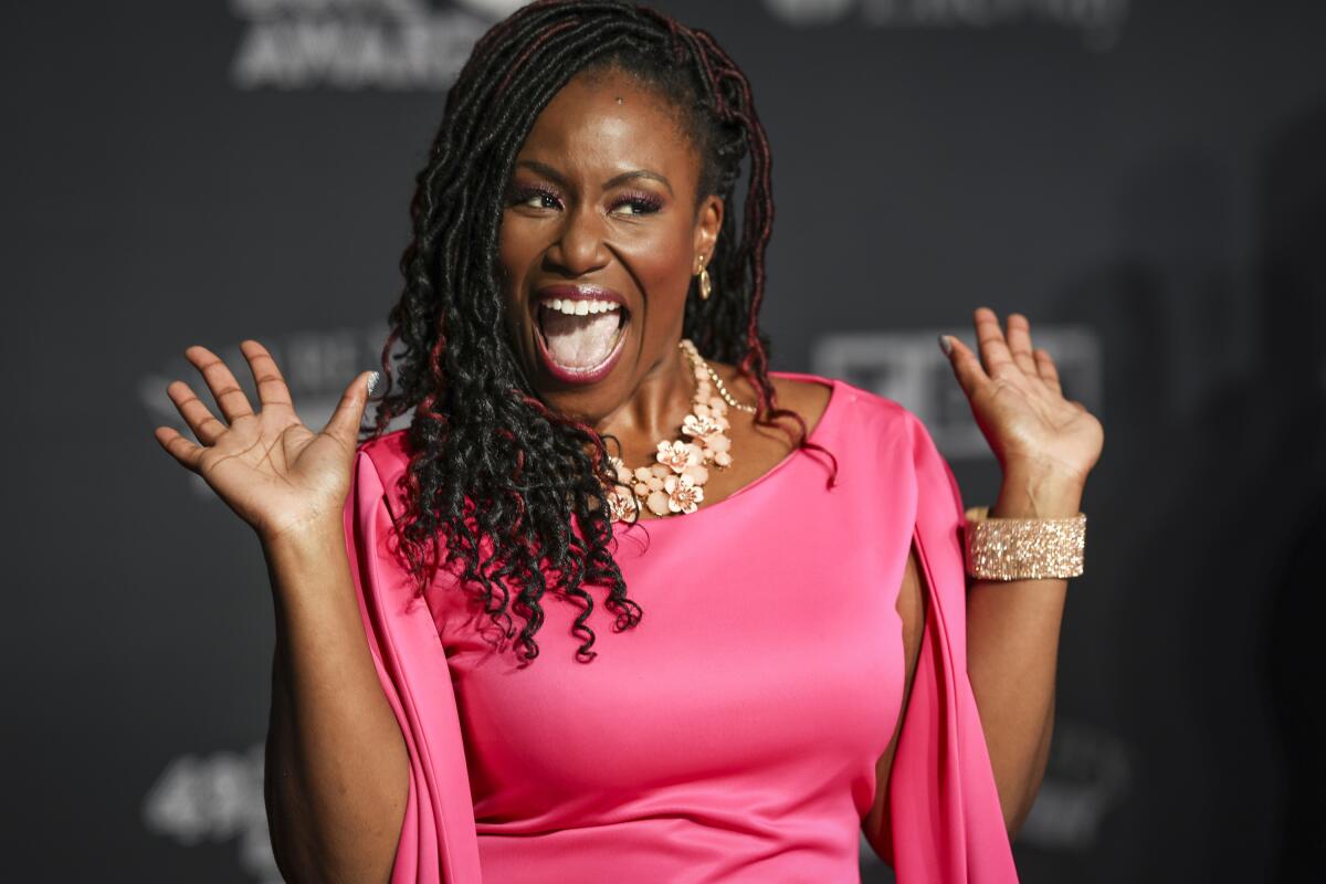 A pink-clad Mandisa holds her hands up with her fingers splayed while smiling in front of a backdrop