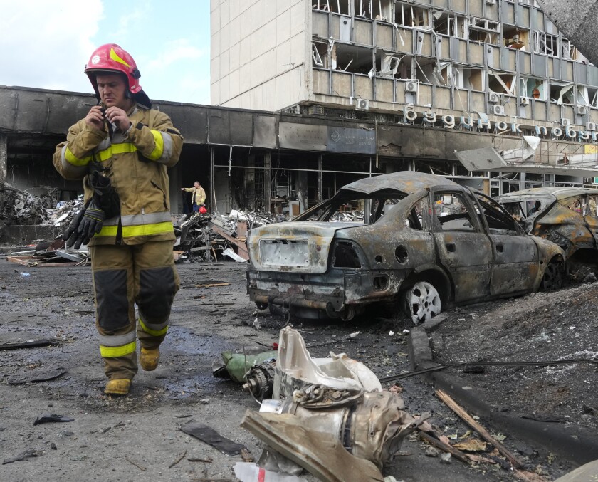 A firefighter stands near burned-out cars and a building with broken windows.  
