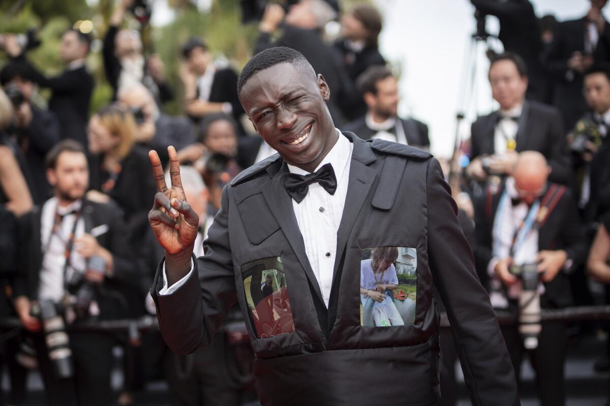 A social media star poses on the red carpet in a black tuxedo, flashing a peace sign as photographers gather behind him