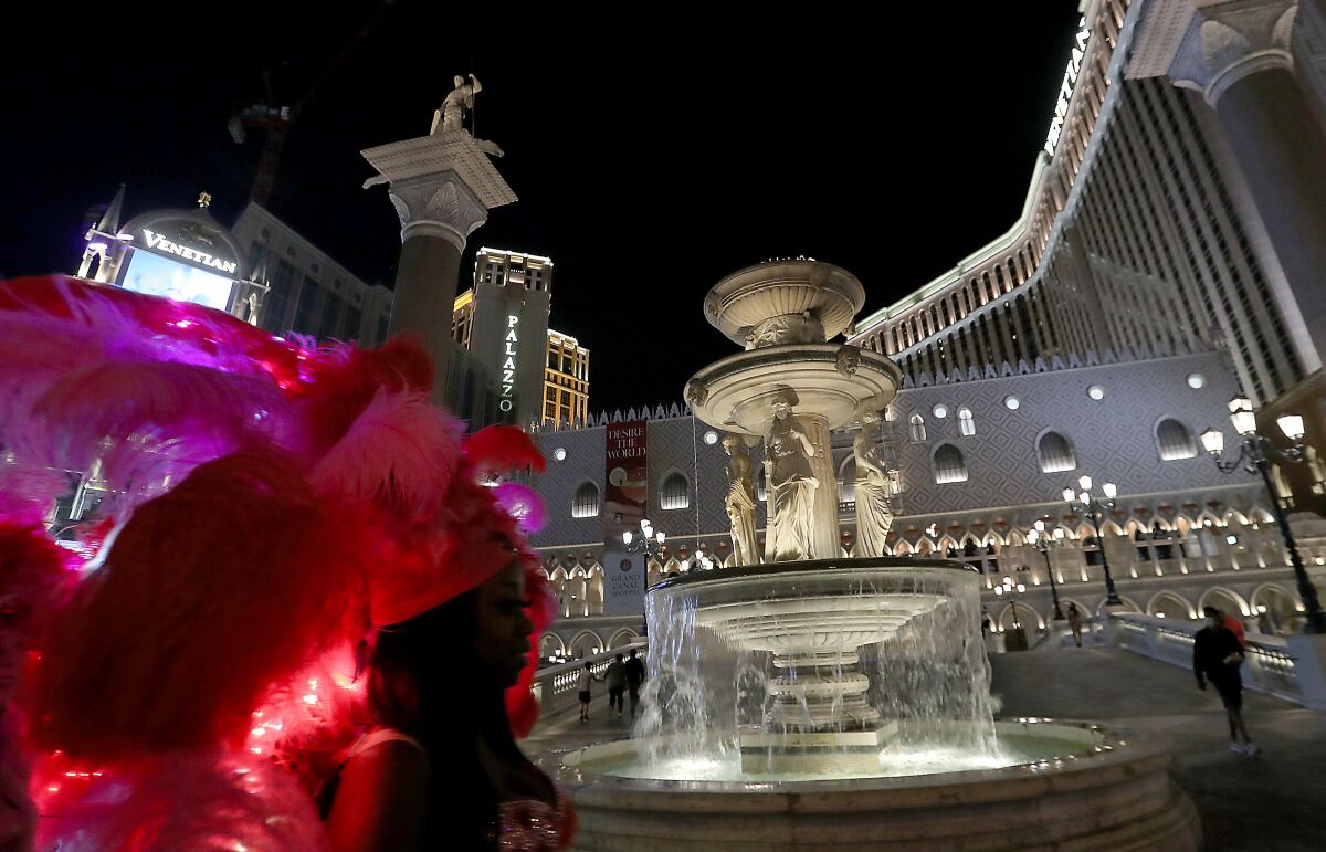 A person in a pink feathered headdress is seen beside an elaborate fountain and a towering hotel at night.