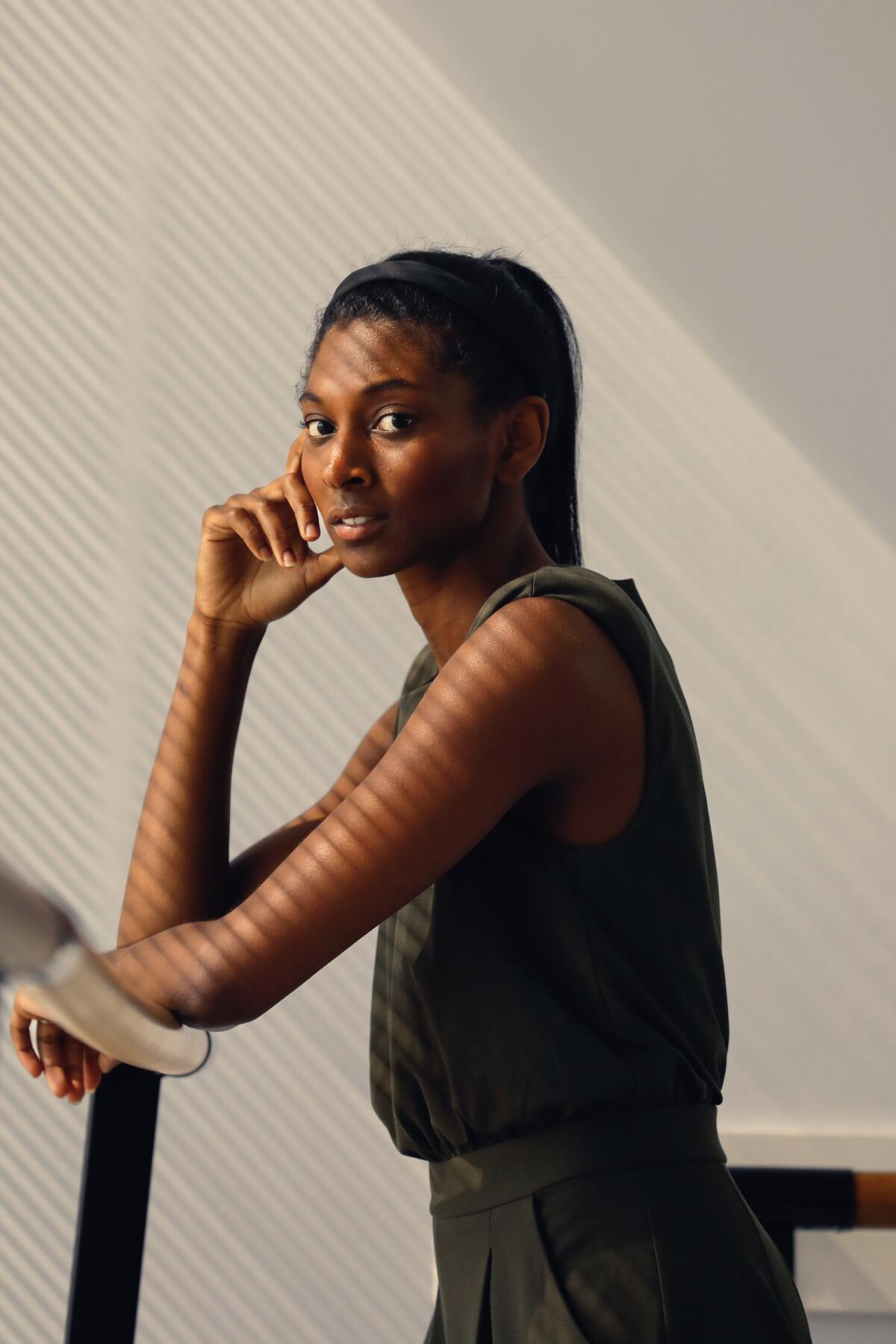 A woman in a sleeveless top stands leaning against a ballet barre, her right hand at her cheek.