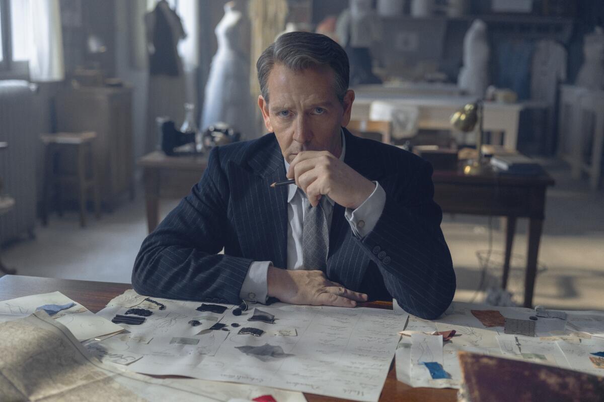 A man in a suit sits at a desk with drawings and holds his hand up to his mouth.