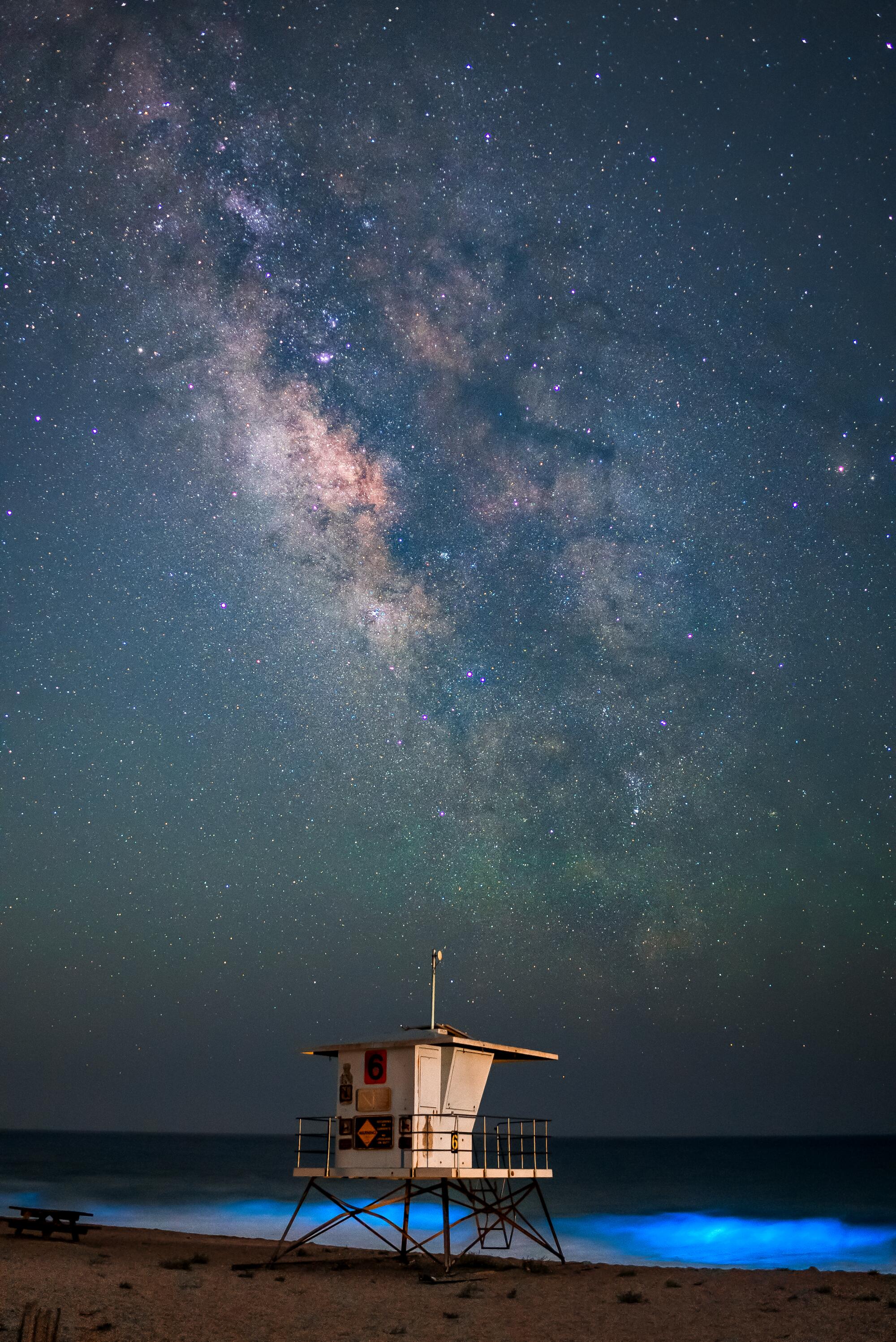 The ocean glows under the Milky Way while a lifeguard station sits on the beach nearby.