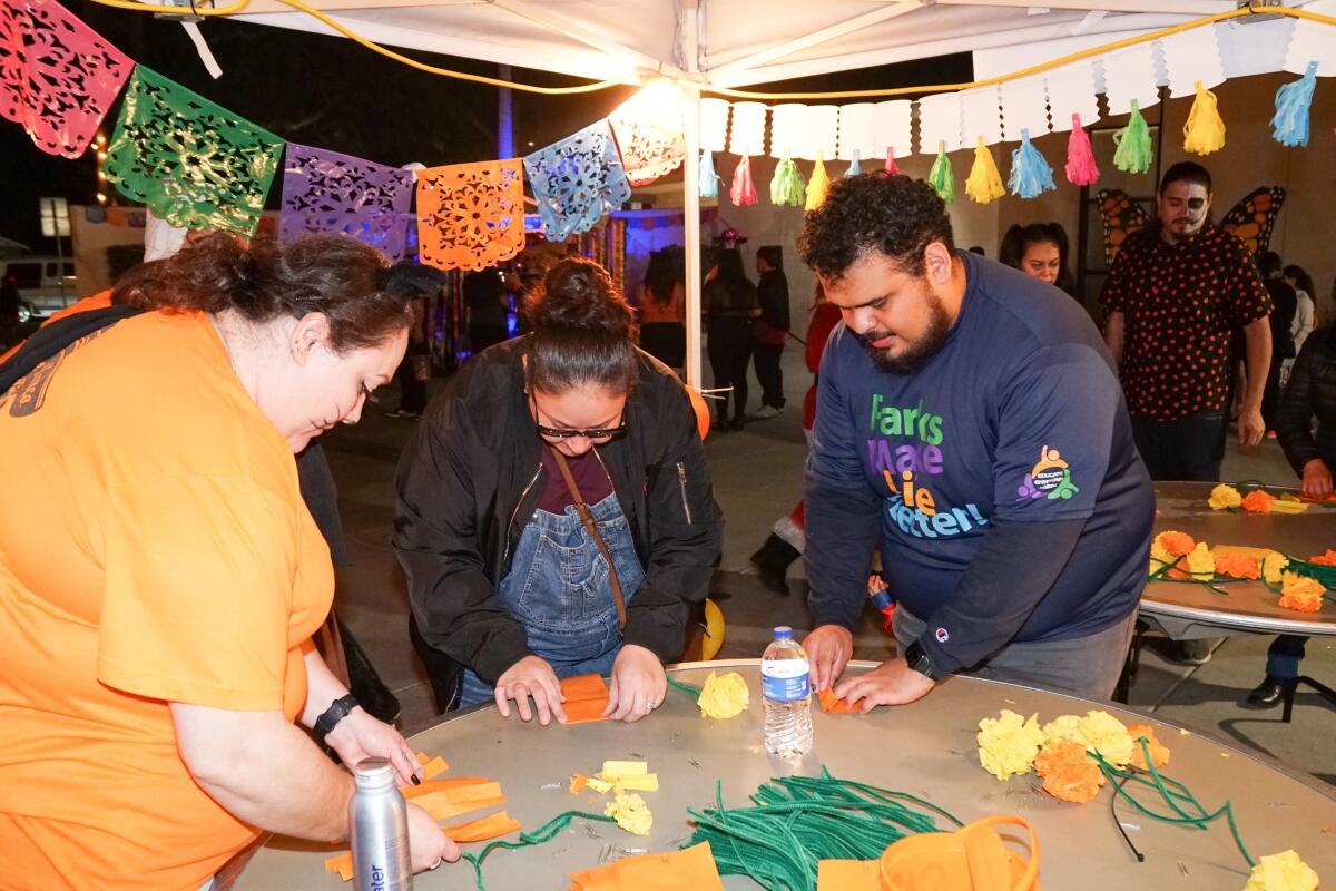 People making paper marigolds stand under strings of papel picado