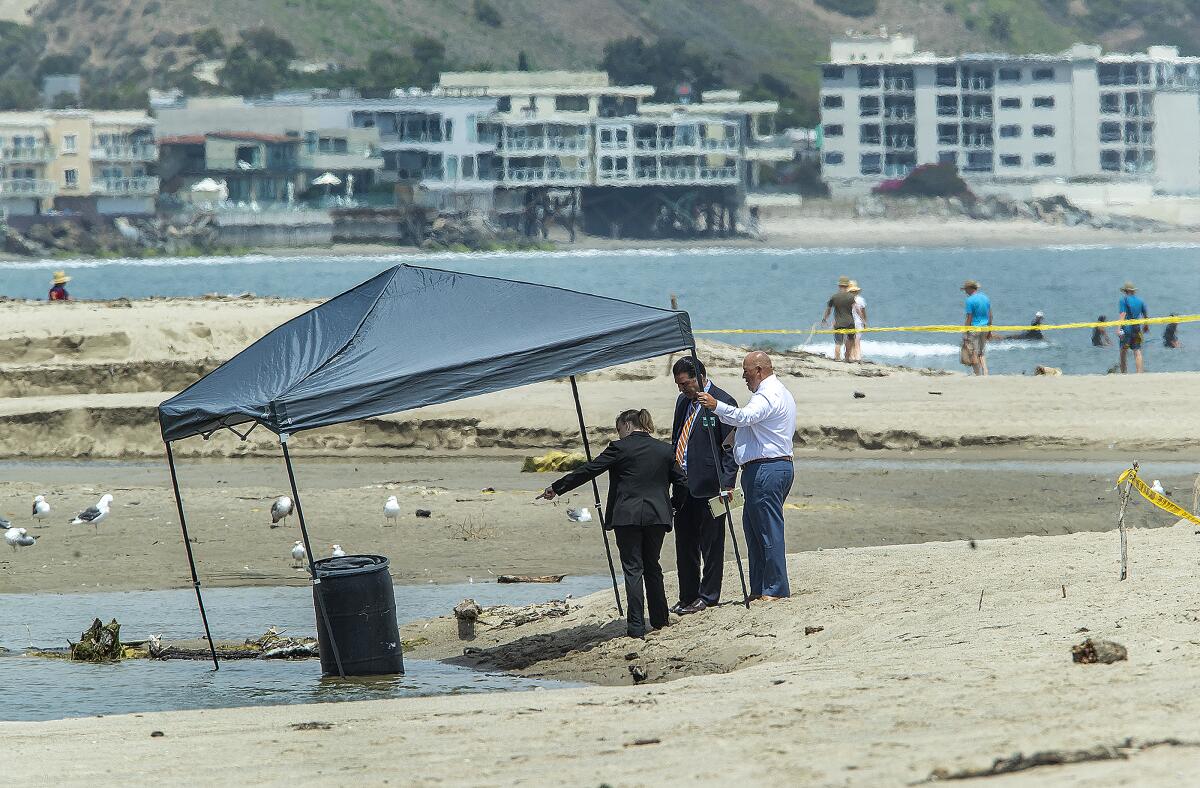 A barrel is seen under a pop-up canopy on a beach. Three people stand nearby pointing.