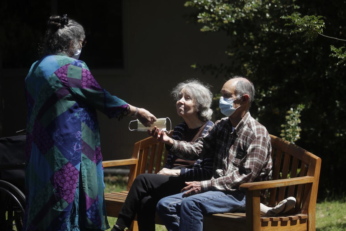 A nursing home director hands a mask to an elderly couple sitting on a bench during an outdoor visit at the facility