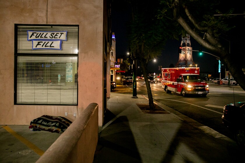 A homeless person sleeps on the ground as an ambulance passes by outside a shuttered nail salon.
