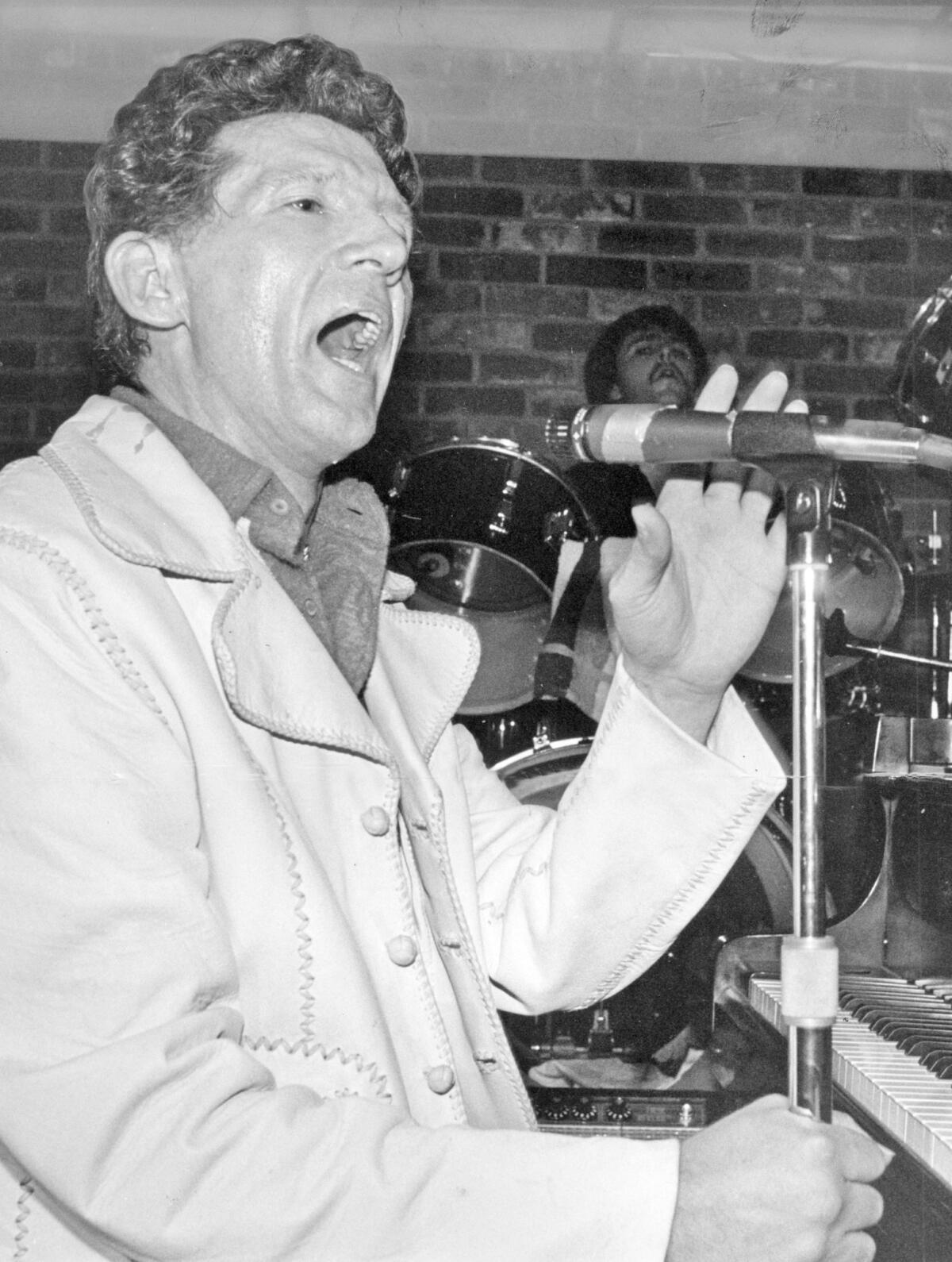 Jerry Lee Lewis, seated at a piano, sings into a microphone. 