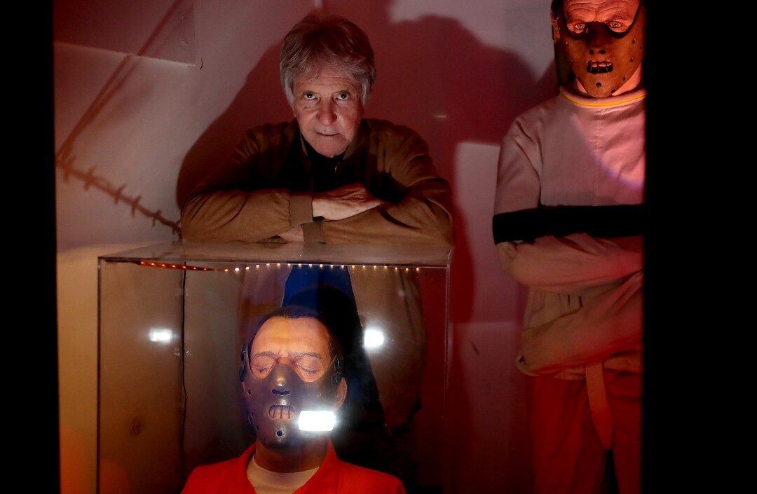 Two men are standing, the man at right wearing a facial mask, as another mask-wearing man sits.