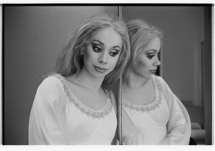In a black and white image, Maria Ewing is seen posing next to a mirror in costume and stage makeup