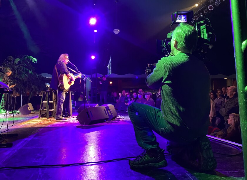 A cameraman records a musician on stage with a guitar on the set of the documentary "The day the music died."