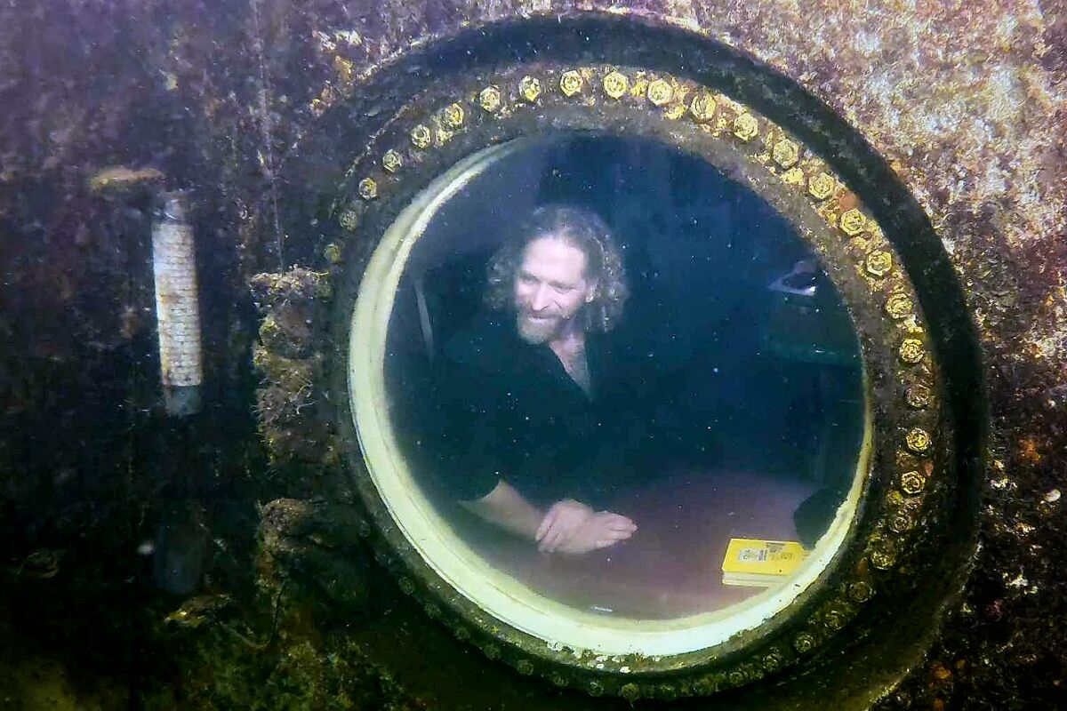 A man looking out of a round porthole from a rusty underwater container