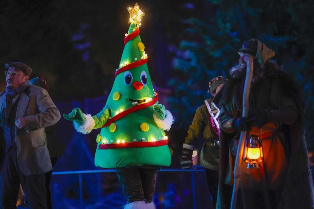 A person in a Christmas tree costume walking around a Christmas village at night.