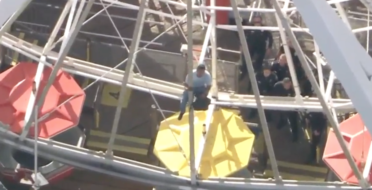 A man sits on the support structure of a Ferris wheel holding a dark bag.
