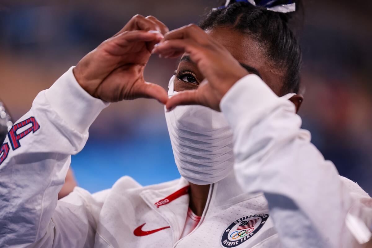 Simone Biles, in a mask, holds up her hands in the shape of a heart.
