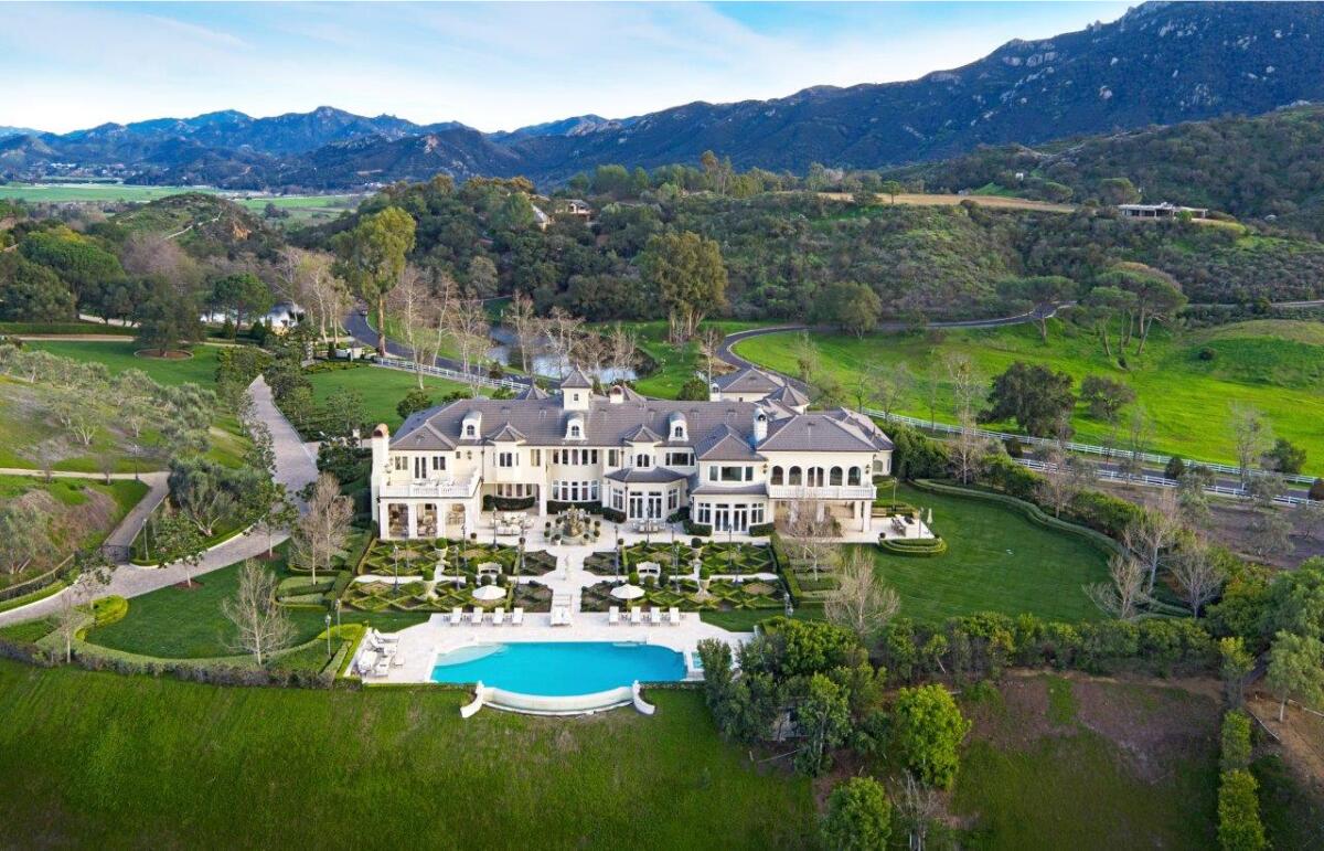 An aerial view shows a mansion and pool amid green lawns and tree-covered hills with mountains as a backdrop.