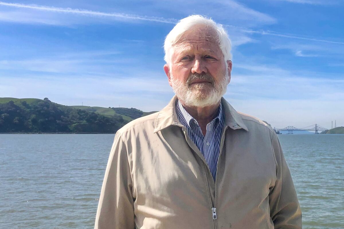 A man with white hair and a beard in front of an expanse of water, with land on the other side and a bridge in the distance.