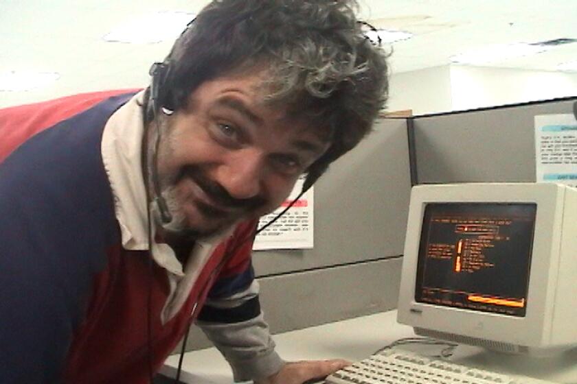 A smiling man wearing a phone headset leans down in front of a 1990s computer monitor.