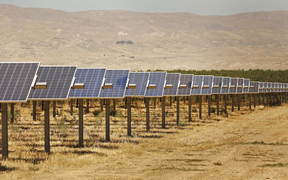 Rows of solar panels are seen on empty acres with low brown hills in the background.