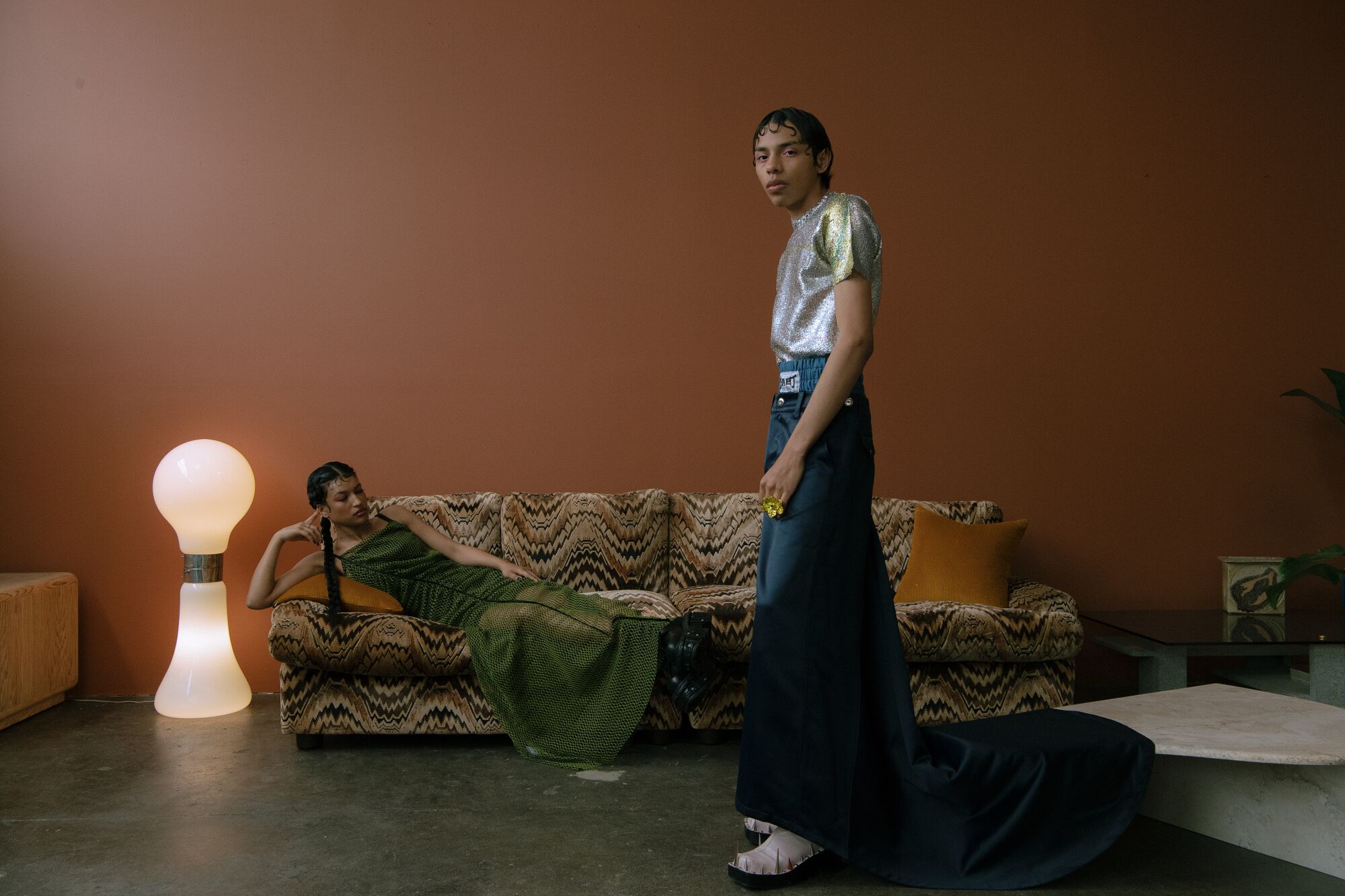 A model in a green dress lies on a couch, while another model stands in a long skirt that drapes over a coffee table.