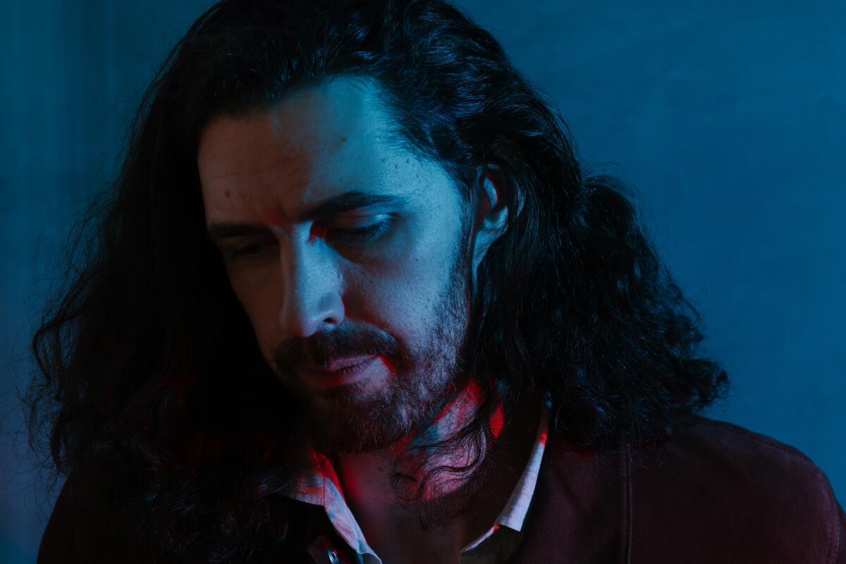 A portrait of a man with long, brown hair looking down, cast in red and blue light.
