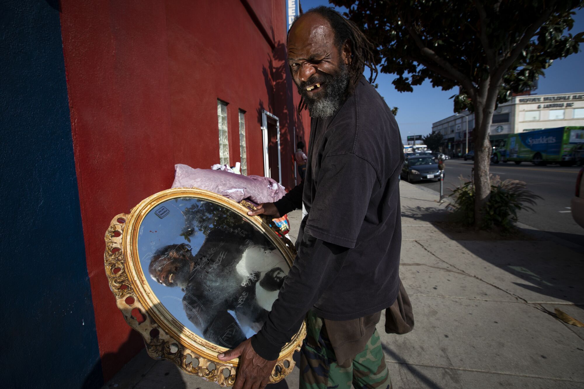 A man stands on a sidewalk holding an ornate mirror in both hands. 