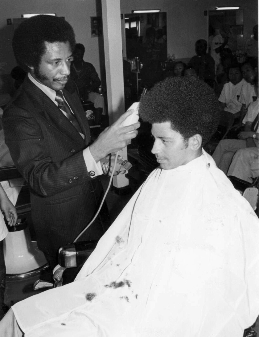 A barber in a suit uses electric clippers on a man with an Afro