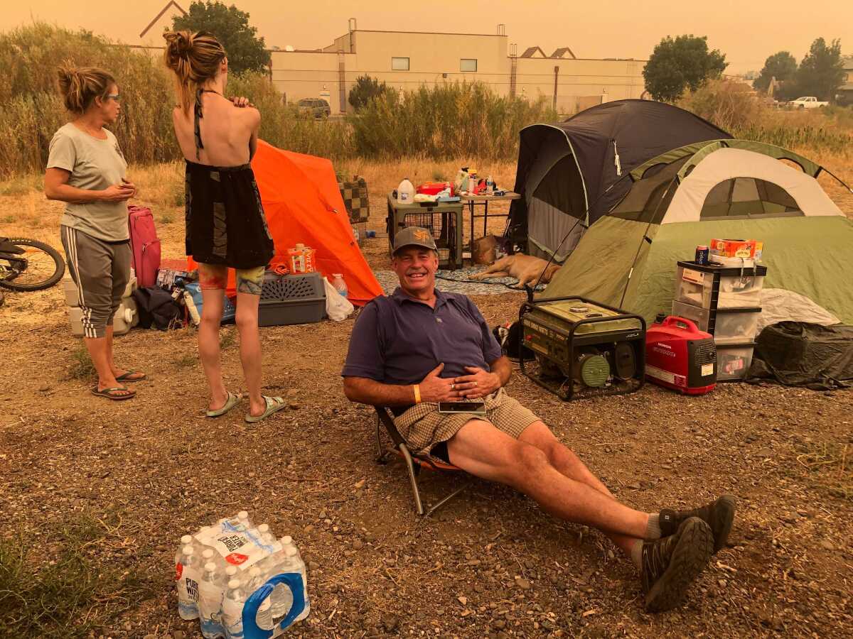 Two people stand and one sits in a low chair amid tents.