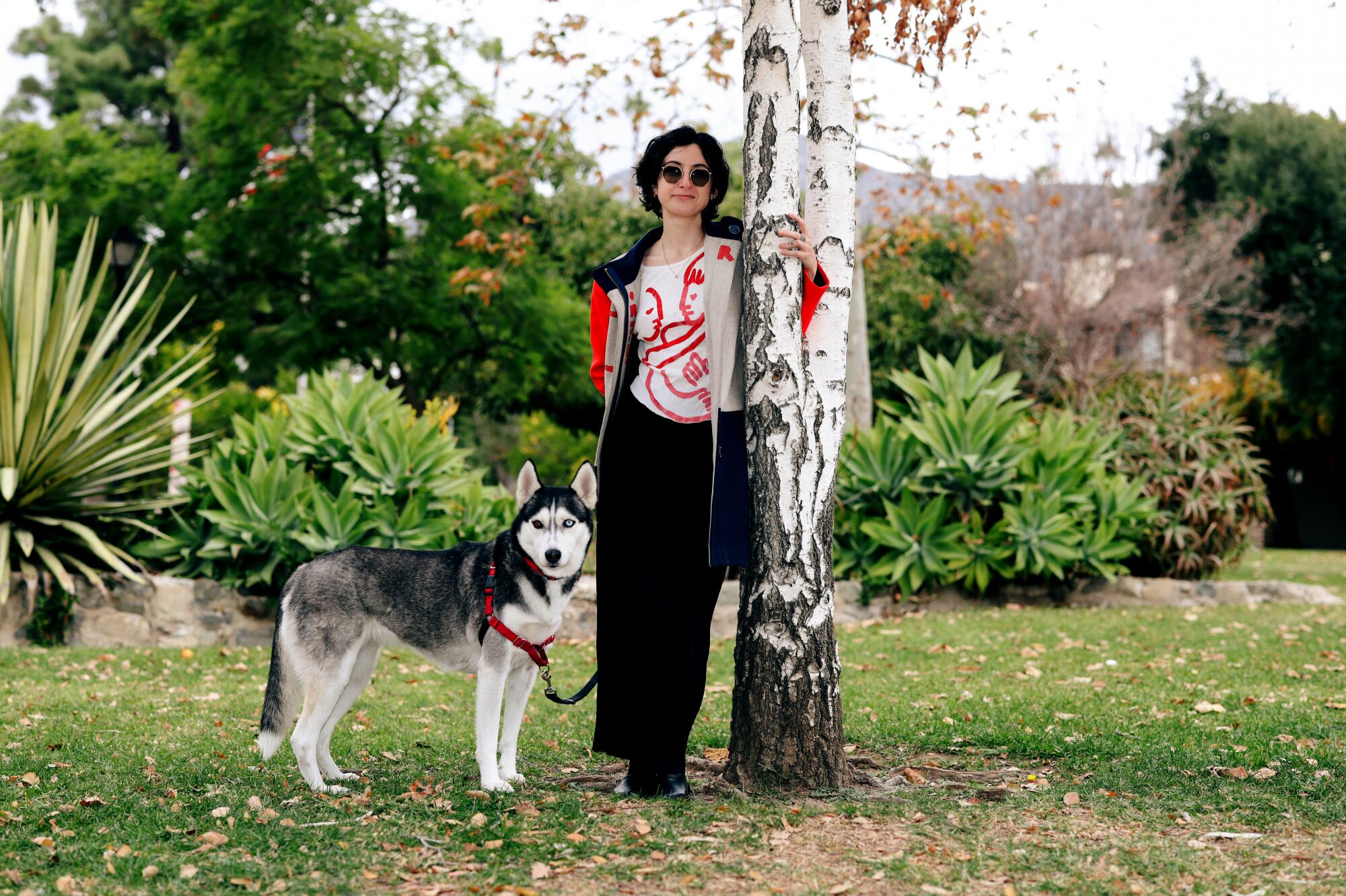 A woman in sunglasses poses next to a tree with her husky.
