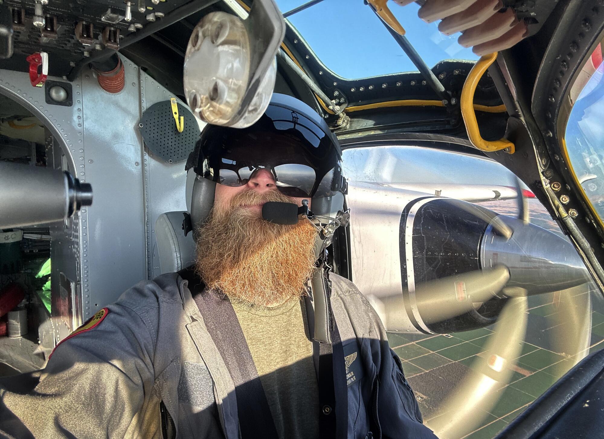 A pilot wearing a helmet and aviator glasses sits in the cockpit of a plane in flight.   