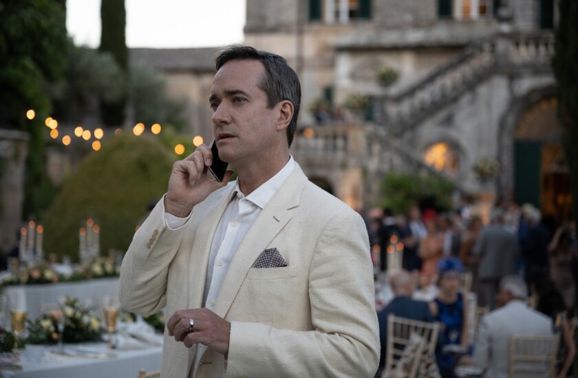 A dapper man in a tan jacket stands away from the crowd at an elegant outdoor reception as he talks on a cellphone.
