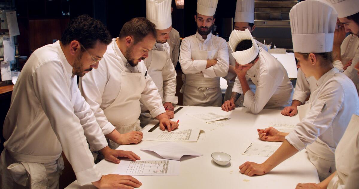 Chefs in white coats and toques surround a table during a meeting.