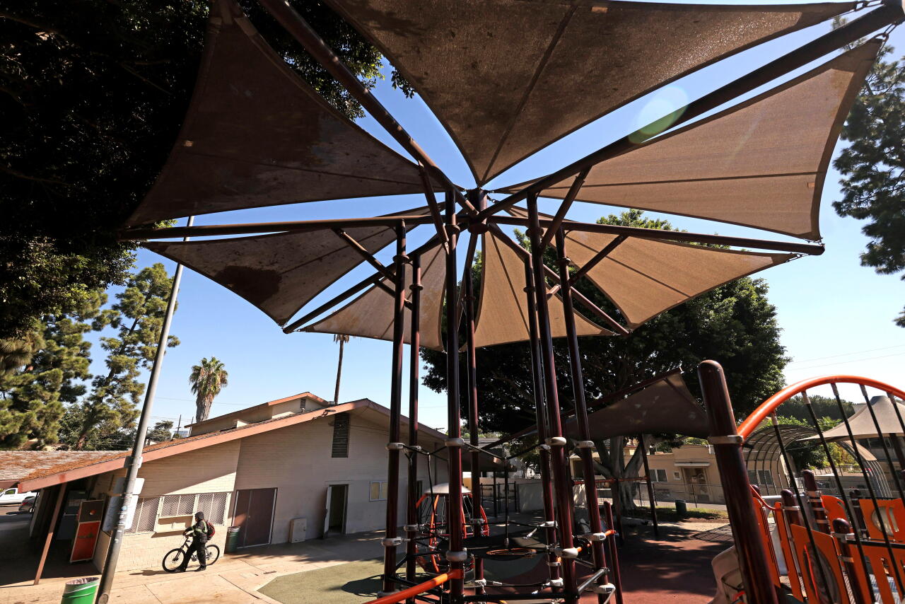 A bicyclist walks near a metal structure with poles and triangular shade sails on top in a playground.
