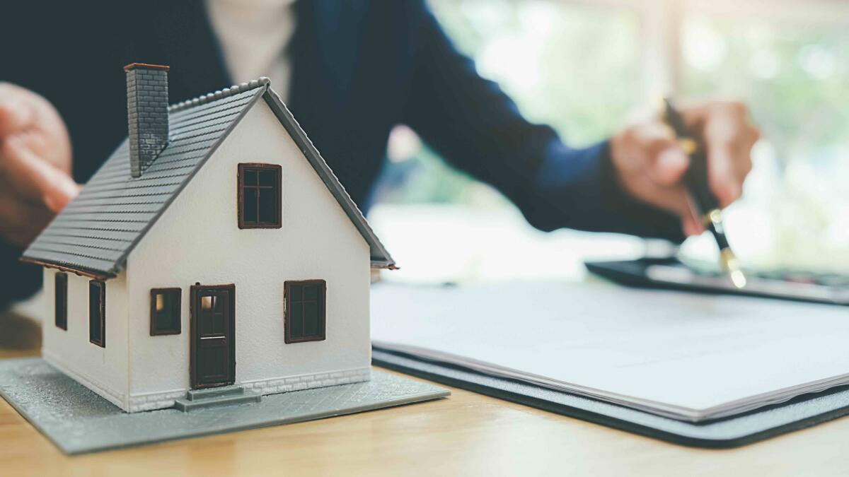 A small house model and documents are on a desk, and an agent's hands point to them both while holding a pen on the left