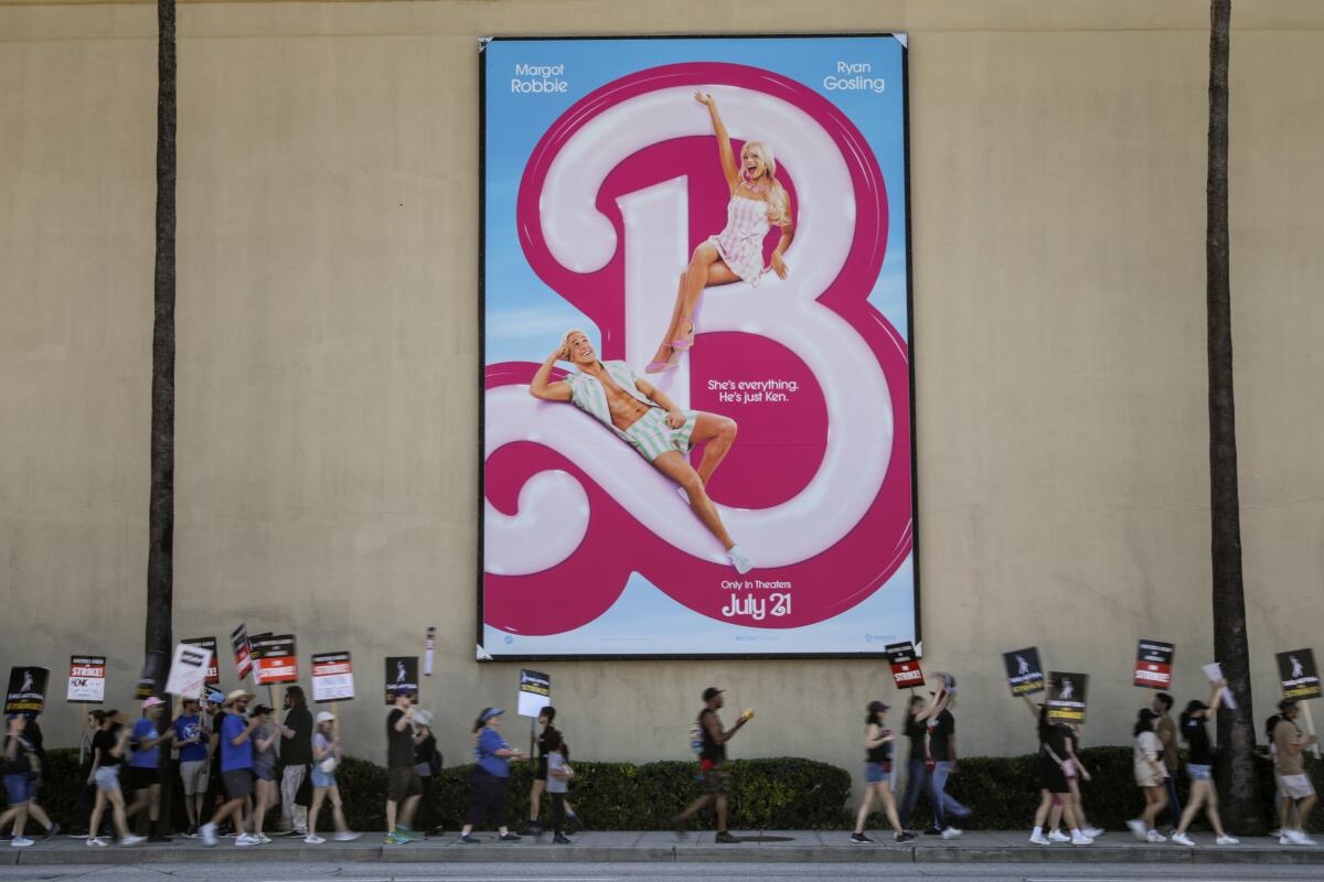 Striking actors and writers with signs walk a picket line below a large ad for Warner Bros' "Barbie" movie.